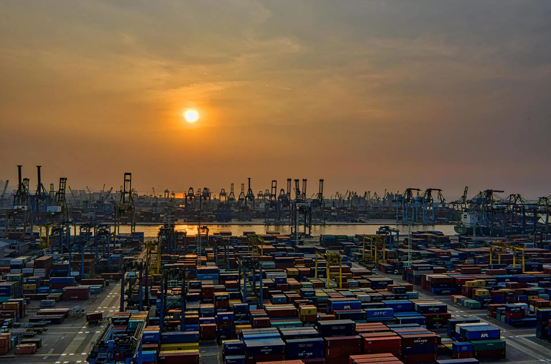 Services Sunset over a bustling seaport filled with stacked shipping containers and cranes.